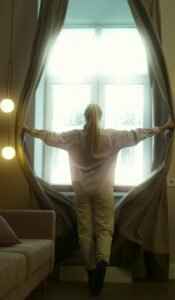 A serene woman opens curtains, welcoming natural light into a cozy room setting.