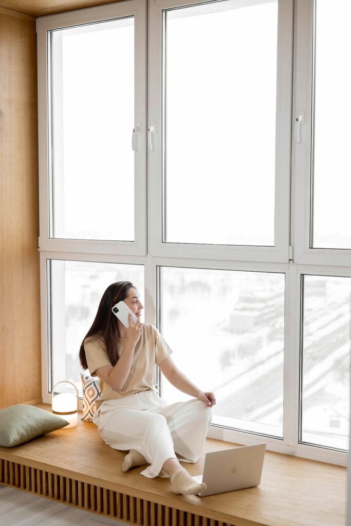 A young woman sits by a large window, talking on the phone with a laptop in front of her.