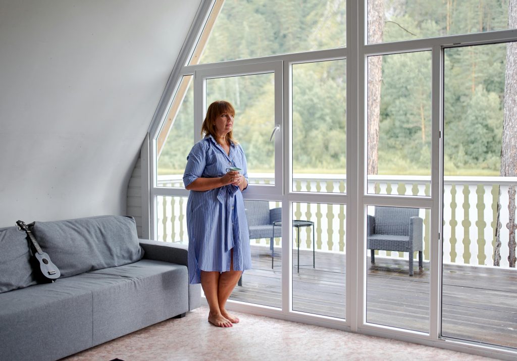 Middle aged barefoot female in casual clothes with cup of beverage looking away near glass wall in room