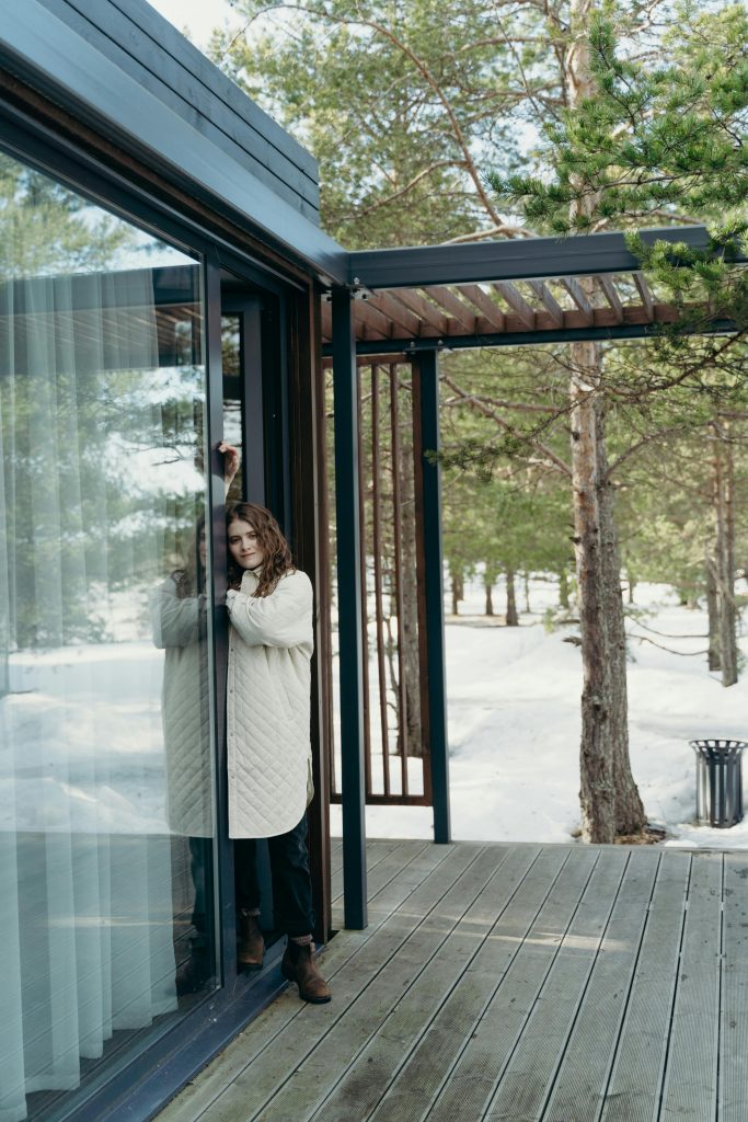 Young woman stands by a glass door in a snowy forest setting.